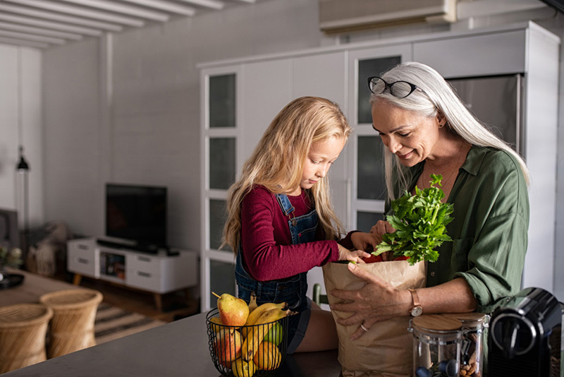 Grandmother and her granddaughter holding grocery shopping bag with vegetables at home. Happy old grandma holding paper bag with vegetables while grandchild removing food from bag. Senior lovely woman and little girl sitting on kitchen counter with groceries.